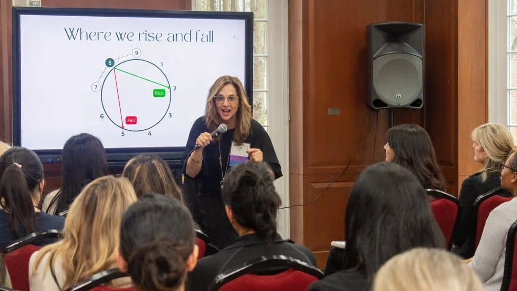 Julie Wright Halbert speaking to a seated audience, holding a microphone beside a screen reading Where we rise and fall